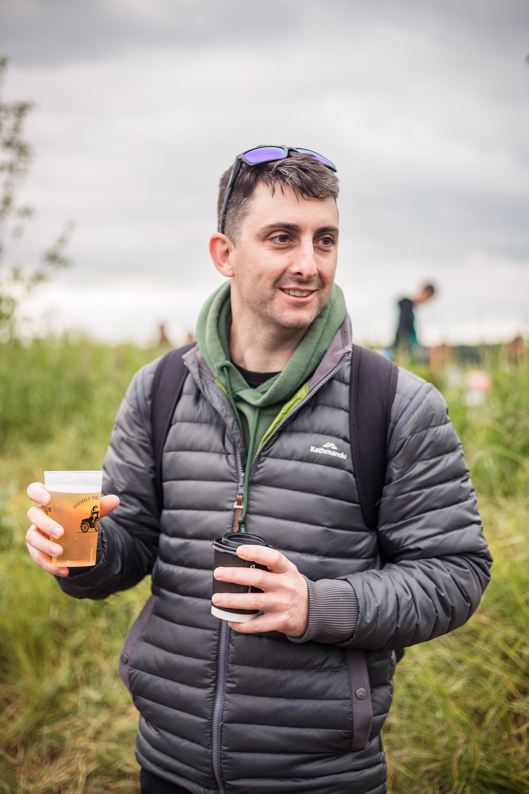 Man wears a coat and backpack holding a beer in the queue at Diddly Squat Farm Shop