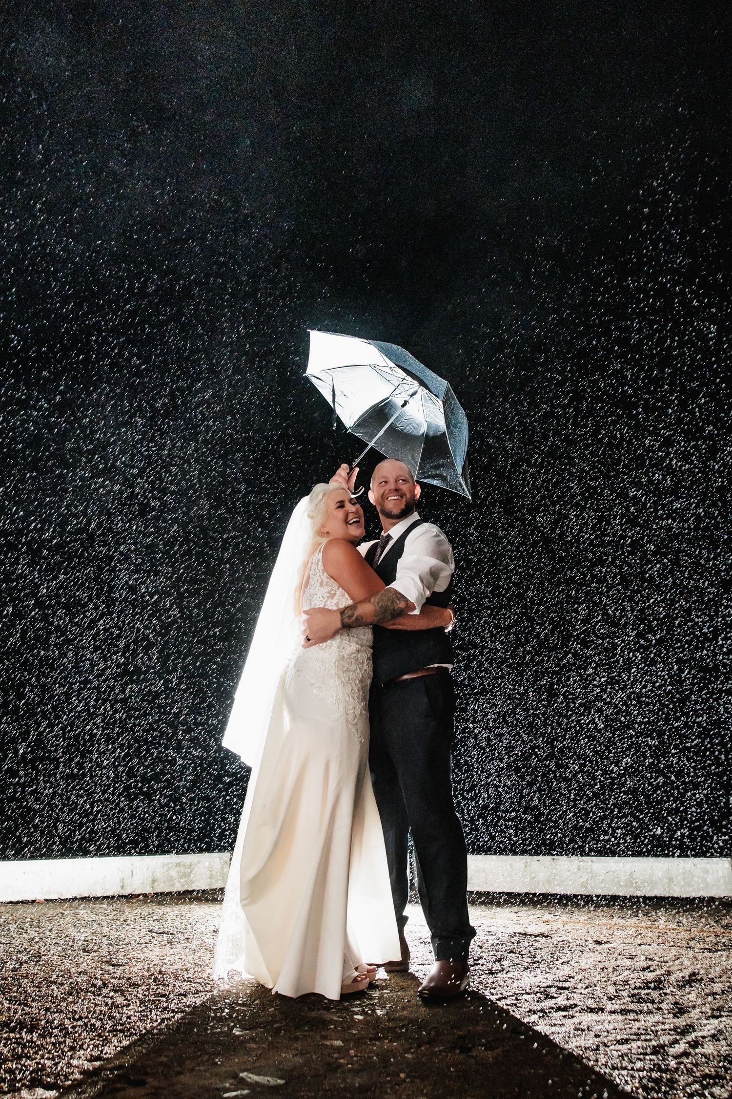 Romantic black and white wedding photo of couple under clear umbrella in natural outdoor setting.