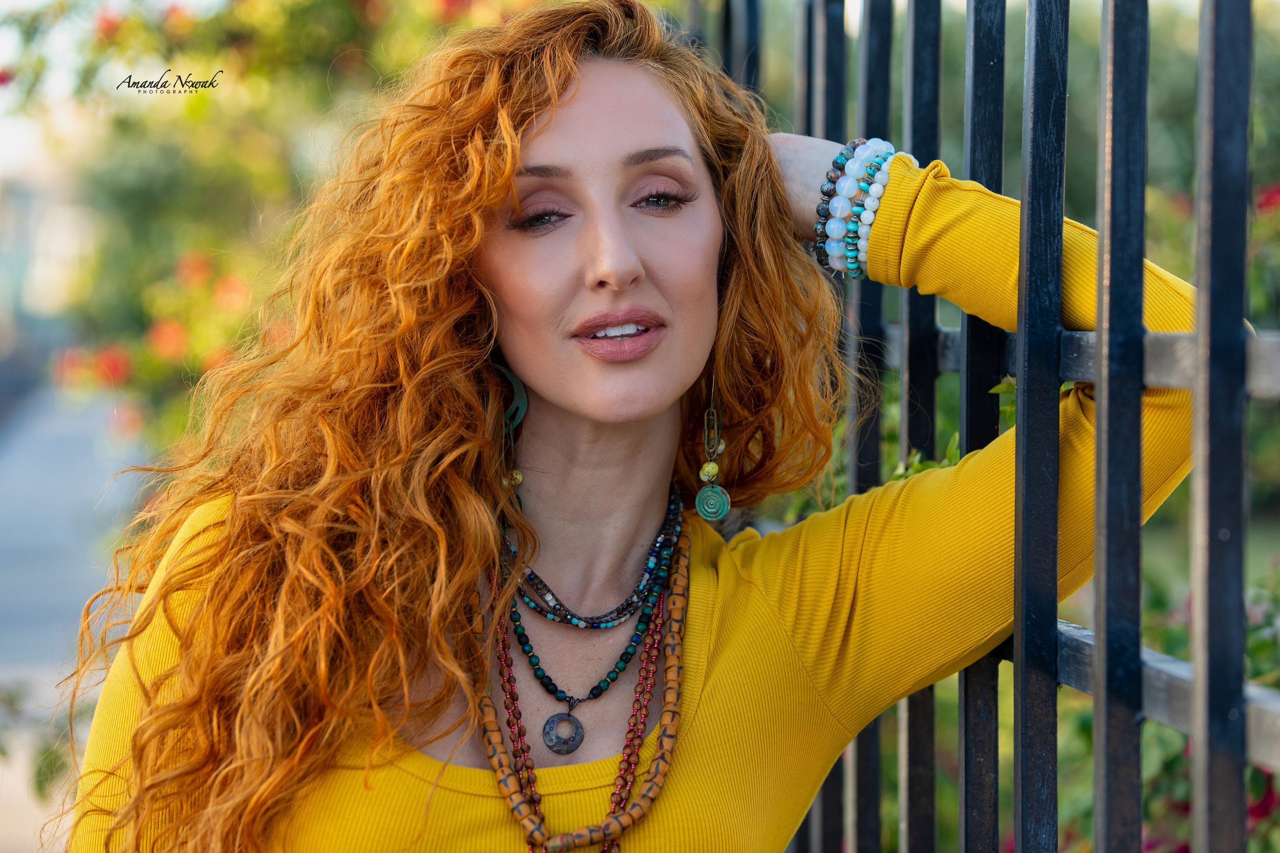 Woman with long curly red hair in yellow top leaning on iron fence outdoors.