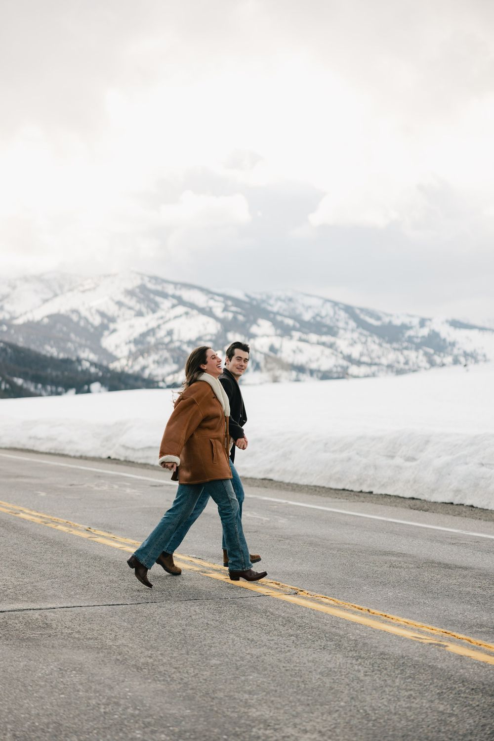 Couple walking on snowy mountain road wearing winter coats.