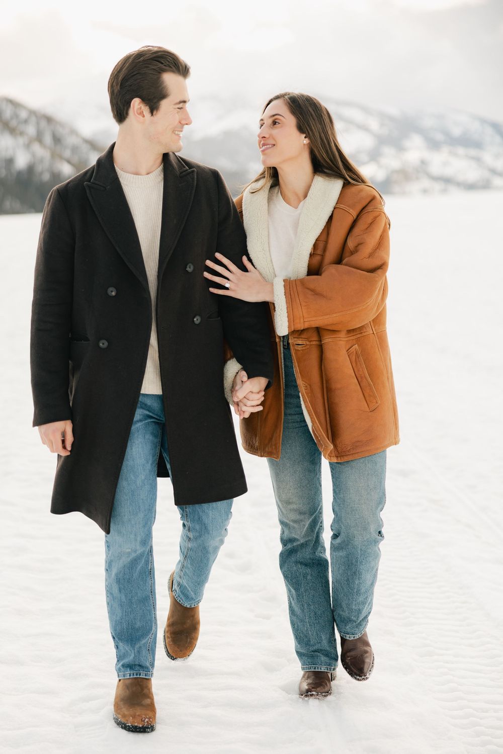 Couple walking together in snow with mountain backdrop wearing winter coats.