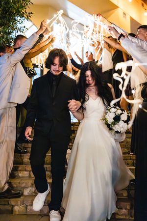 Newlywed couple walks down steps under sparklers held by guests, bride in white gown holding bouquet, groom in black suit.