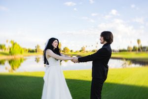 Bride and groom holding hands outdoors near a pond on a sunny day at a wedding celebration.