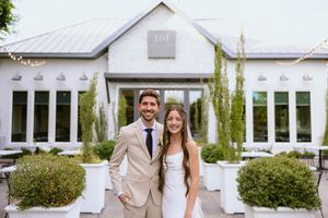 A smiling bride and groom pose together in front of a white wedding venue with lush greenery and string lights.