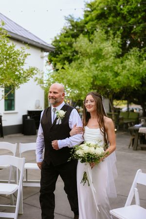Father walks bride down the aisle at outdoor wedding ceremony, bride holds white bouquet.