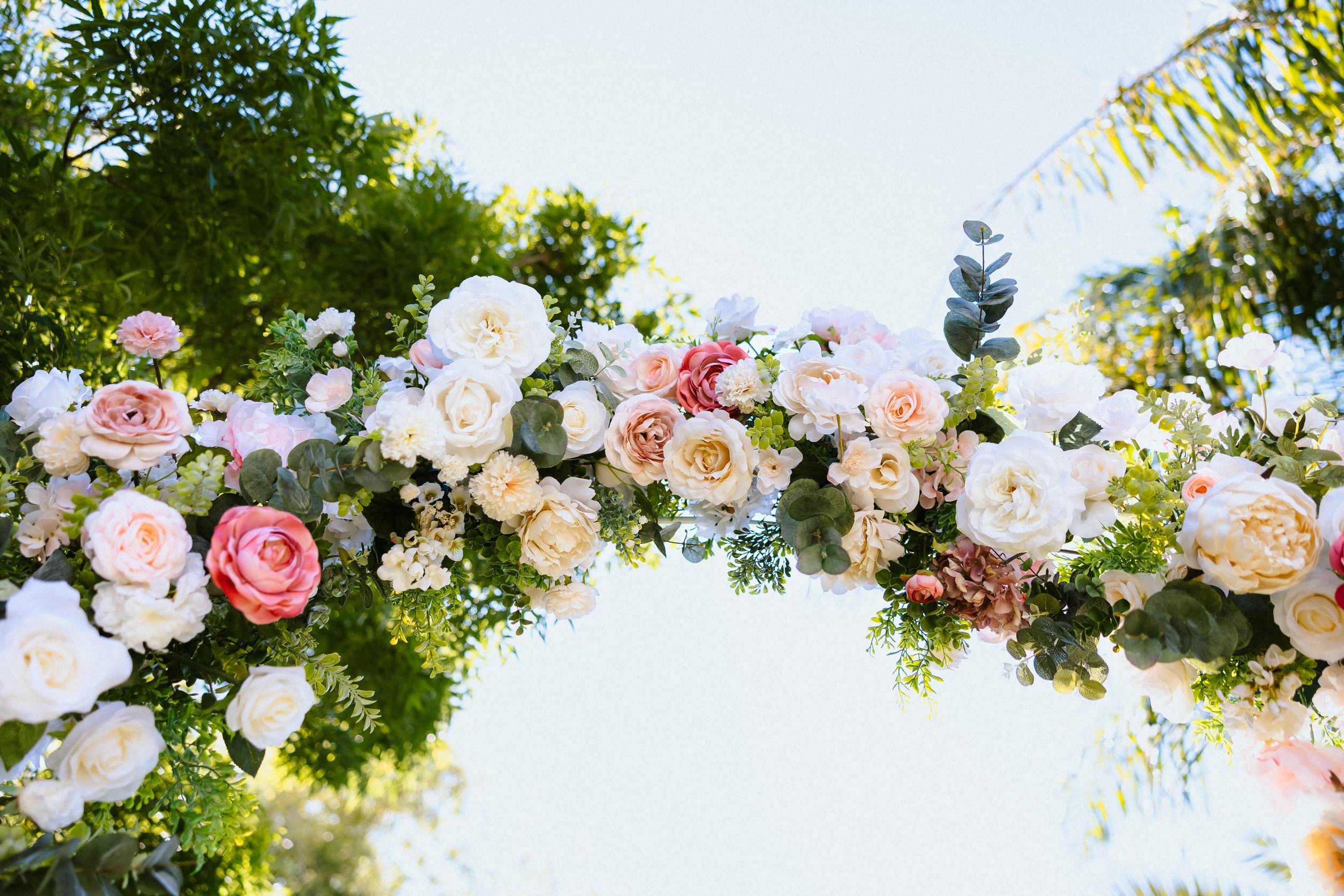 Elegant outdoor wedding arch adorned with blush pink, white, and peach roses surrounded by lush greenery.