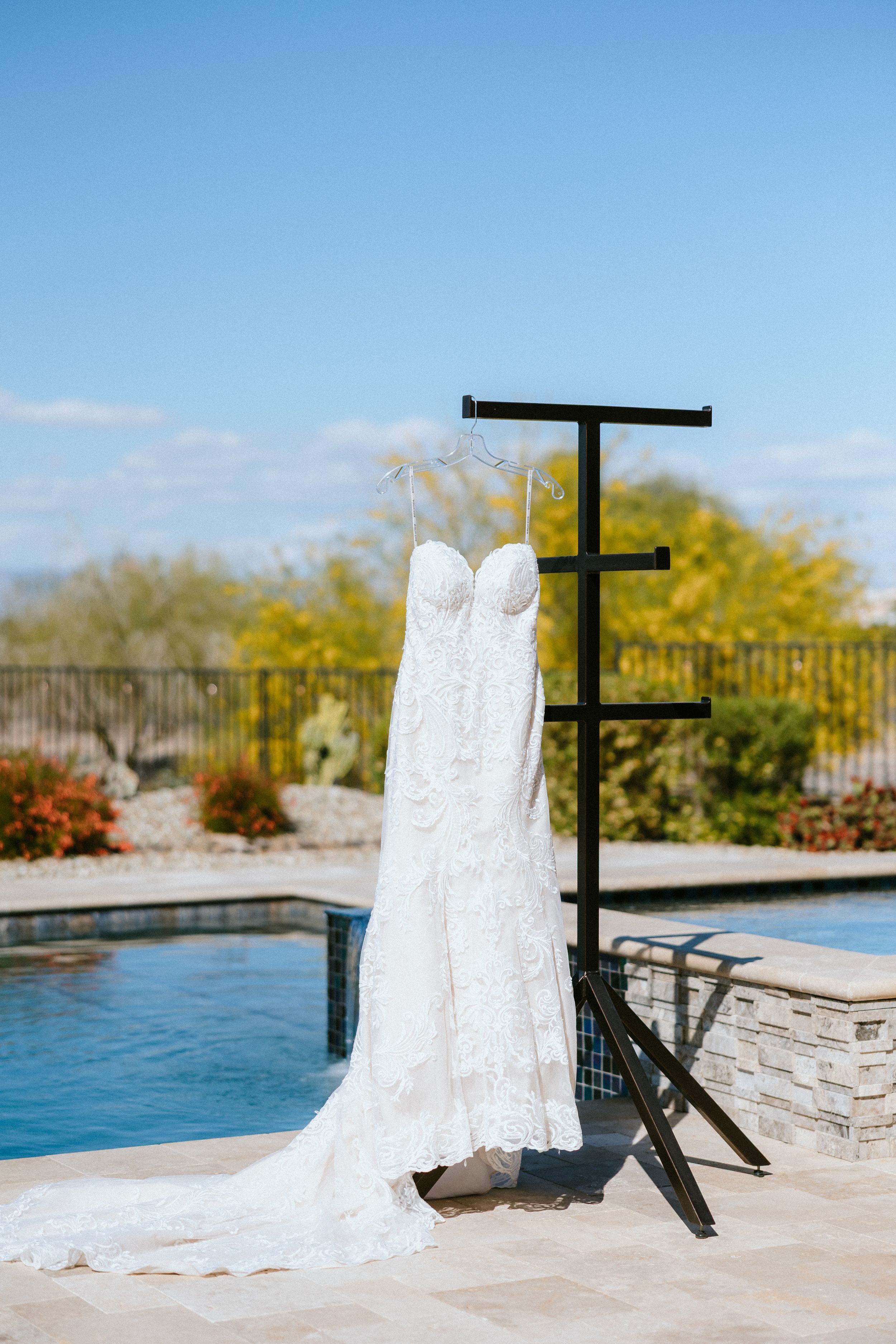 White lace wedding dress hanging on a stand poolside with desert landscaping and blue sky in the background.