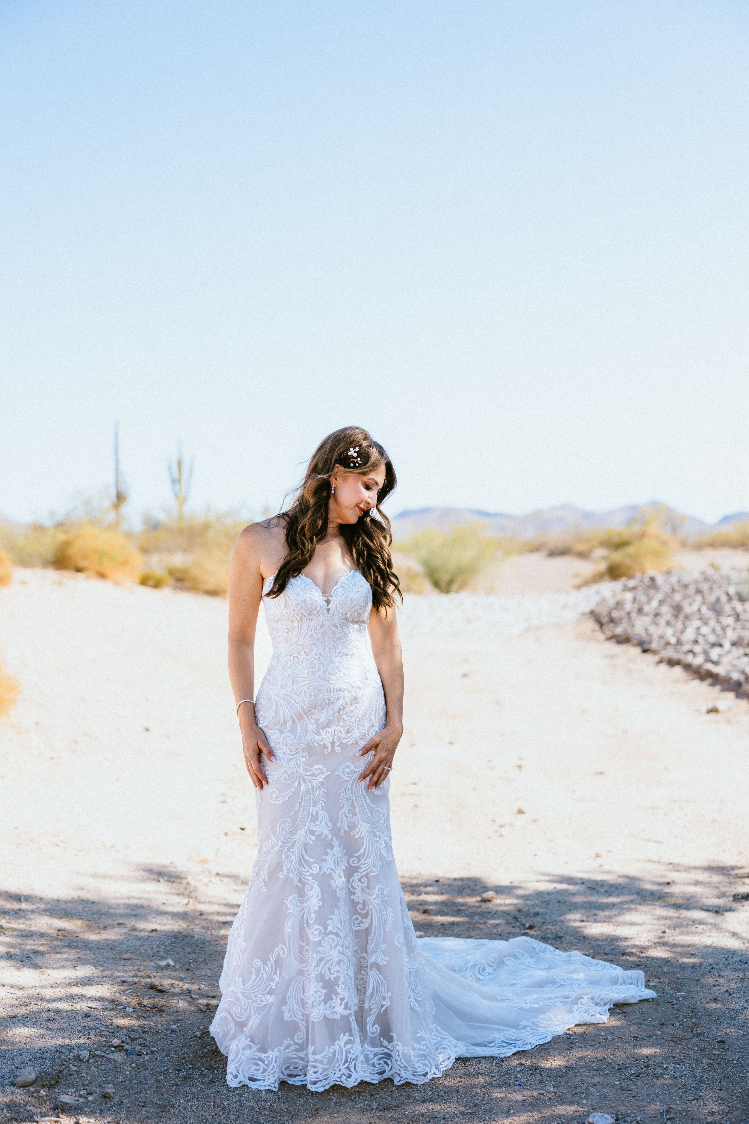 Bride in lace mermaid wedding gown standing on desert path with saguaro cacti and clear blue sky.