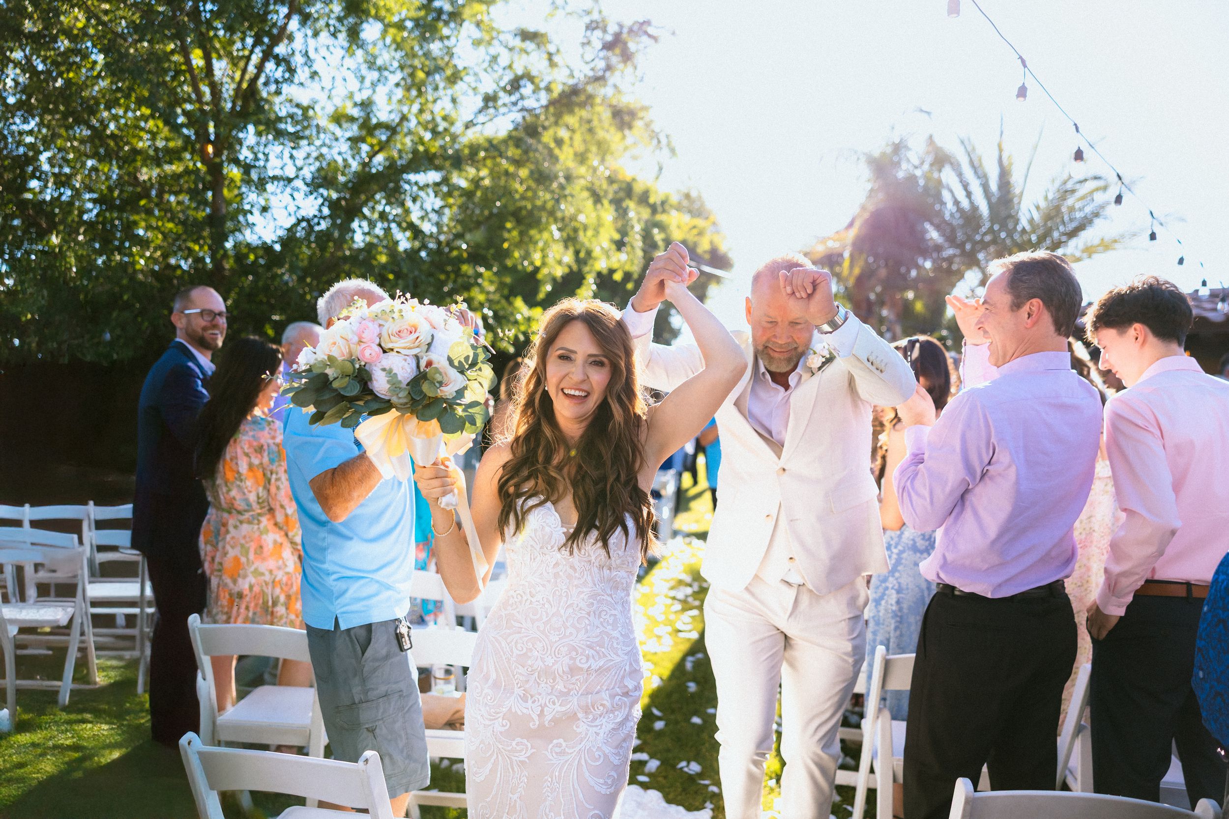 Joyful bride and groom walk down outdoor aisle after wedding ceremony, guests cheering, lush green trees in background.