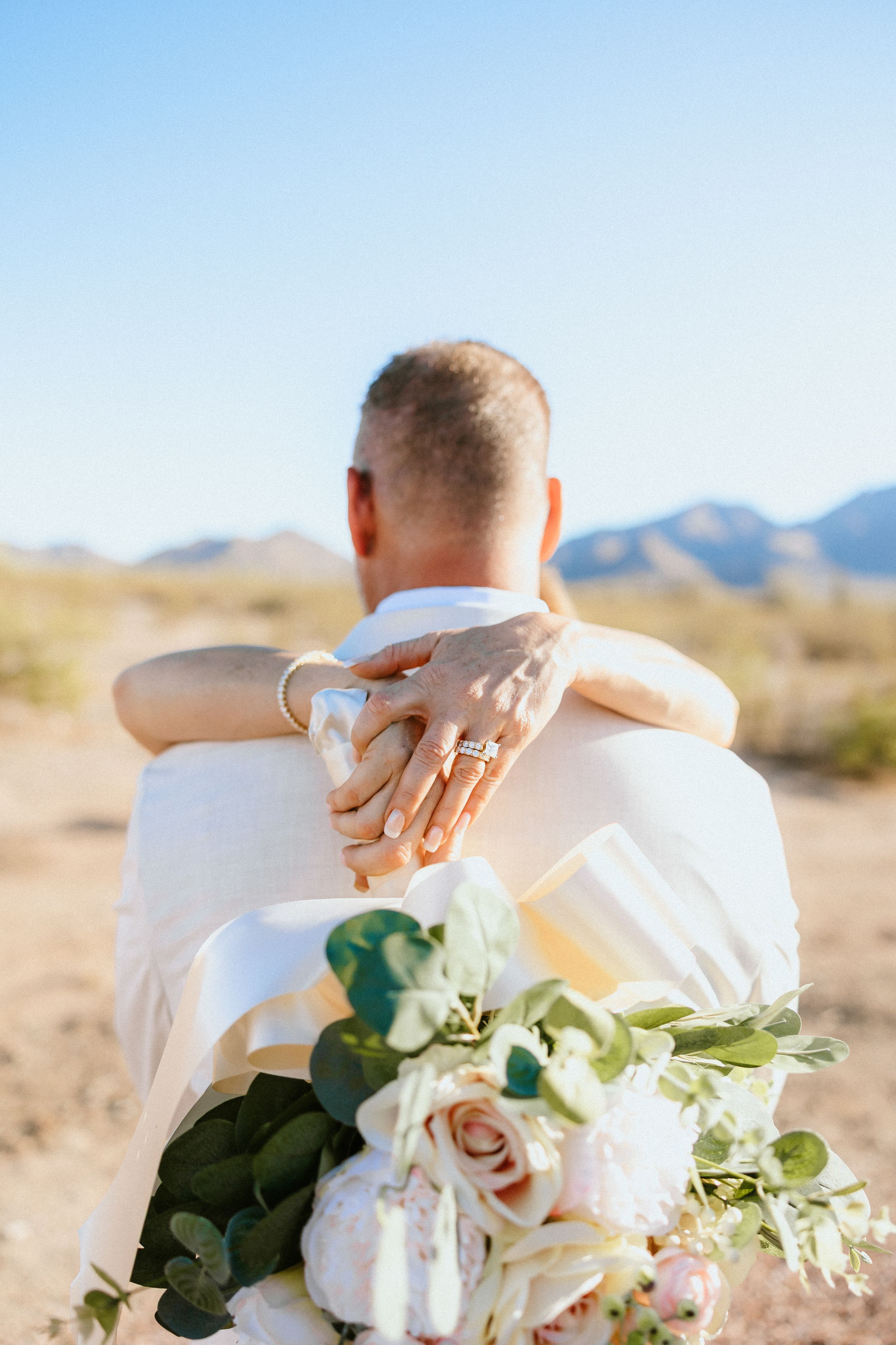 Bride and groom embracing in desert landscape, showcasing wedding rings and bridal bouquet with pink roses.
