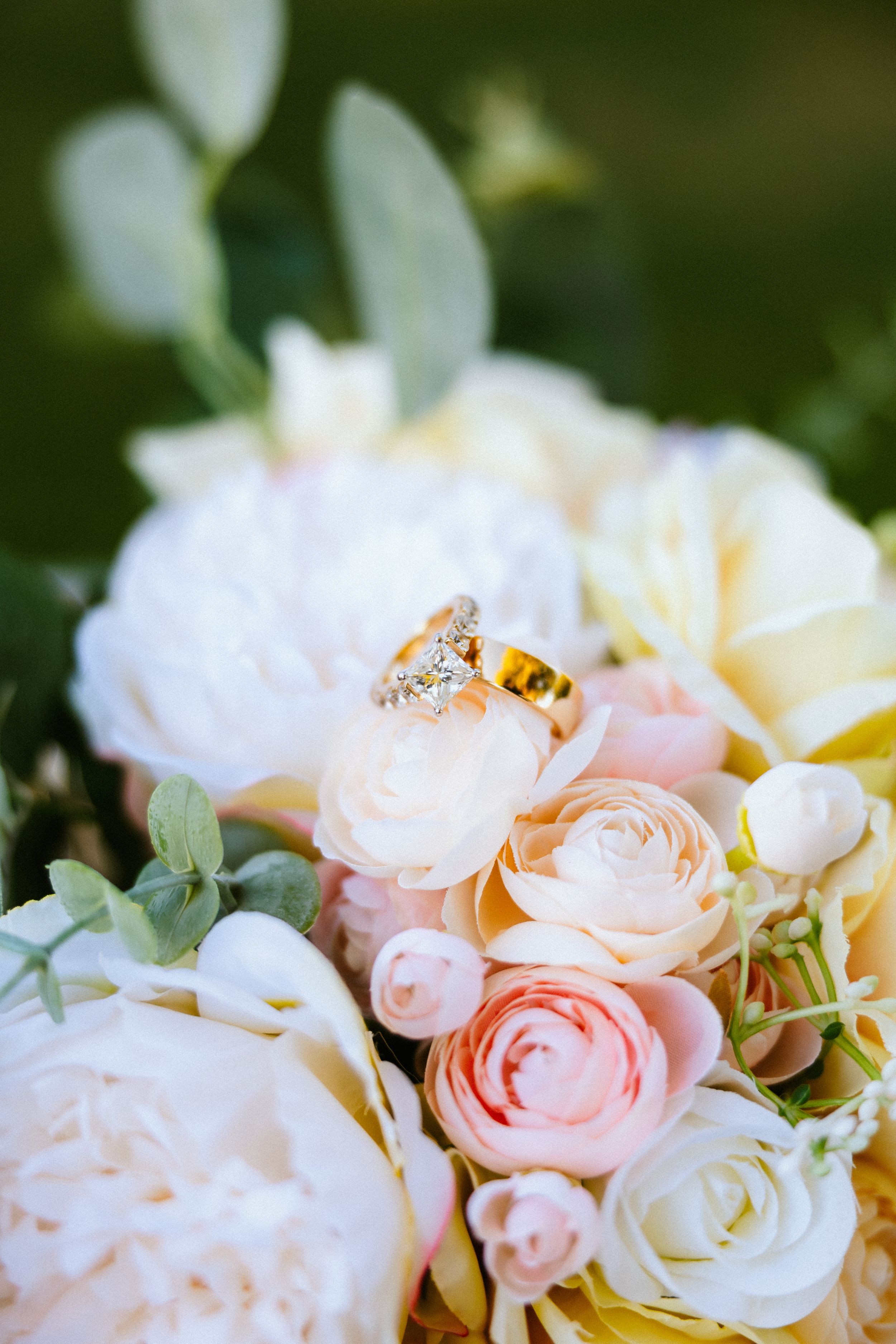 Gold diamond engagement ring resting on a bridal bouquet of soft pink, white, and peach roses and peonies.