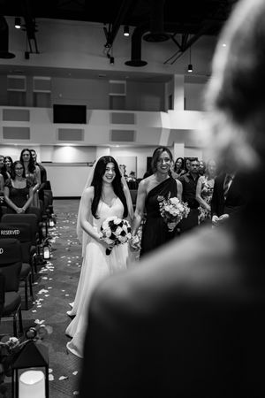 Black and white photo of a smiling bride walking down the aisle with her bridesmaid during a wedding ceremony.
