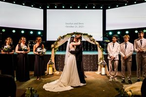 Bride and groom share first kiss at elegant wedding ceremony with floral arch, bridal party, and personalized screen backdrop.