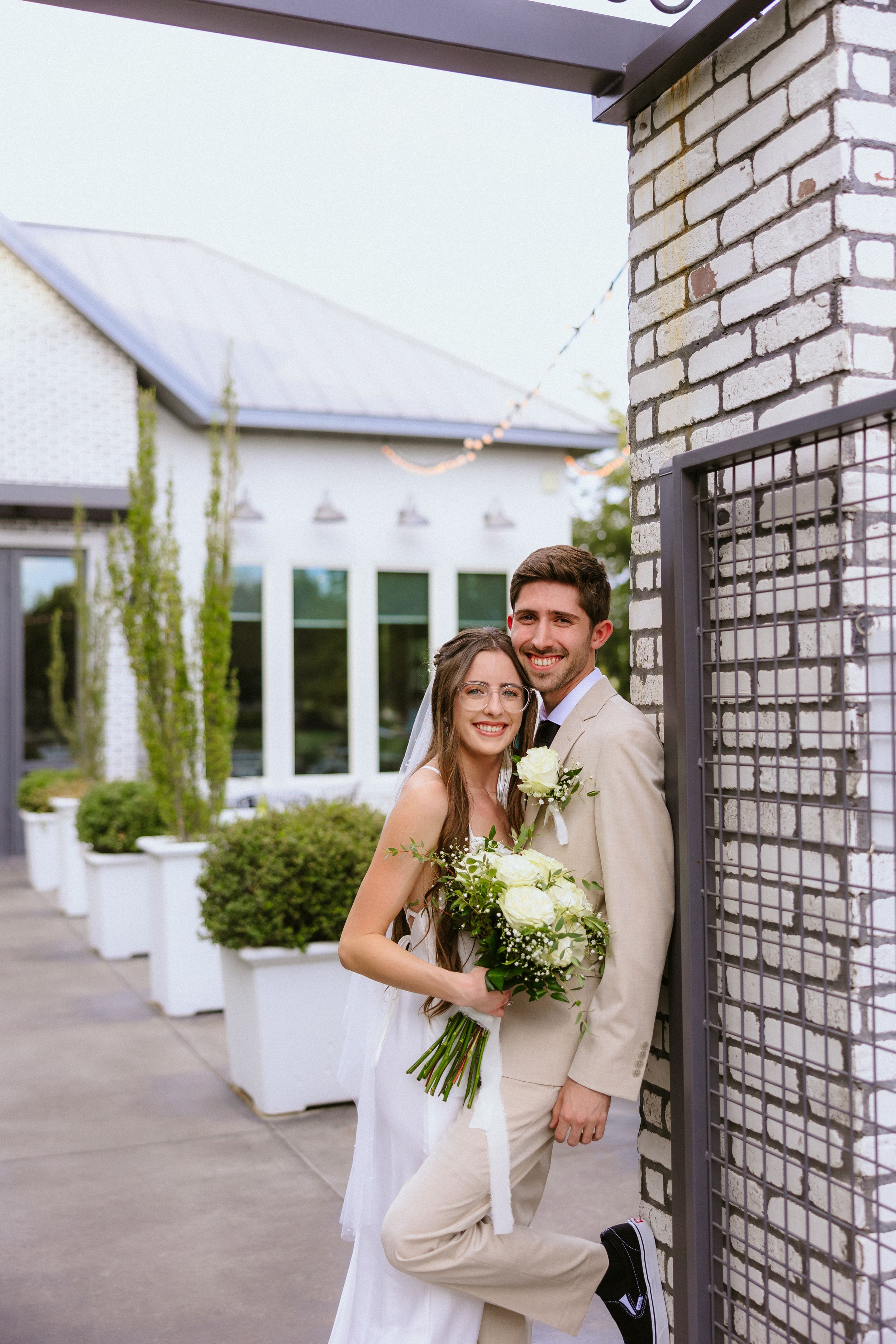 Smiling bride and groom pose together outdoors near brick pillar, bride holding white bouquet, groom in tan suit.