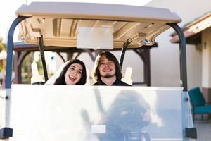 Two young adults smiling and riding together in a white golf cart outdoors.