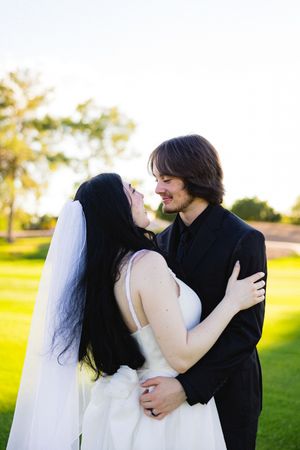 A bride and groom share a romantic embrace outdoors on a sunny day, smiling at each other on a lush green lawn.