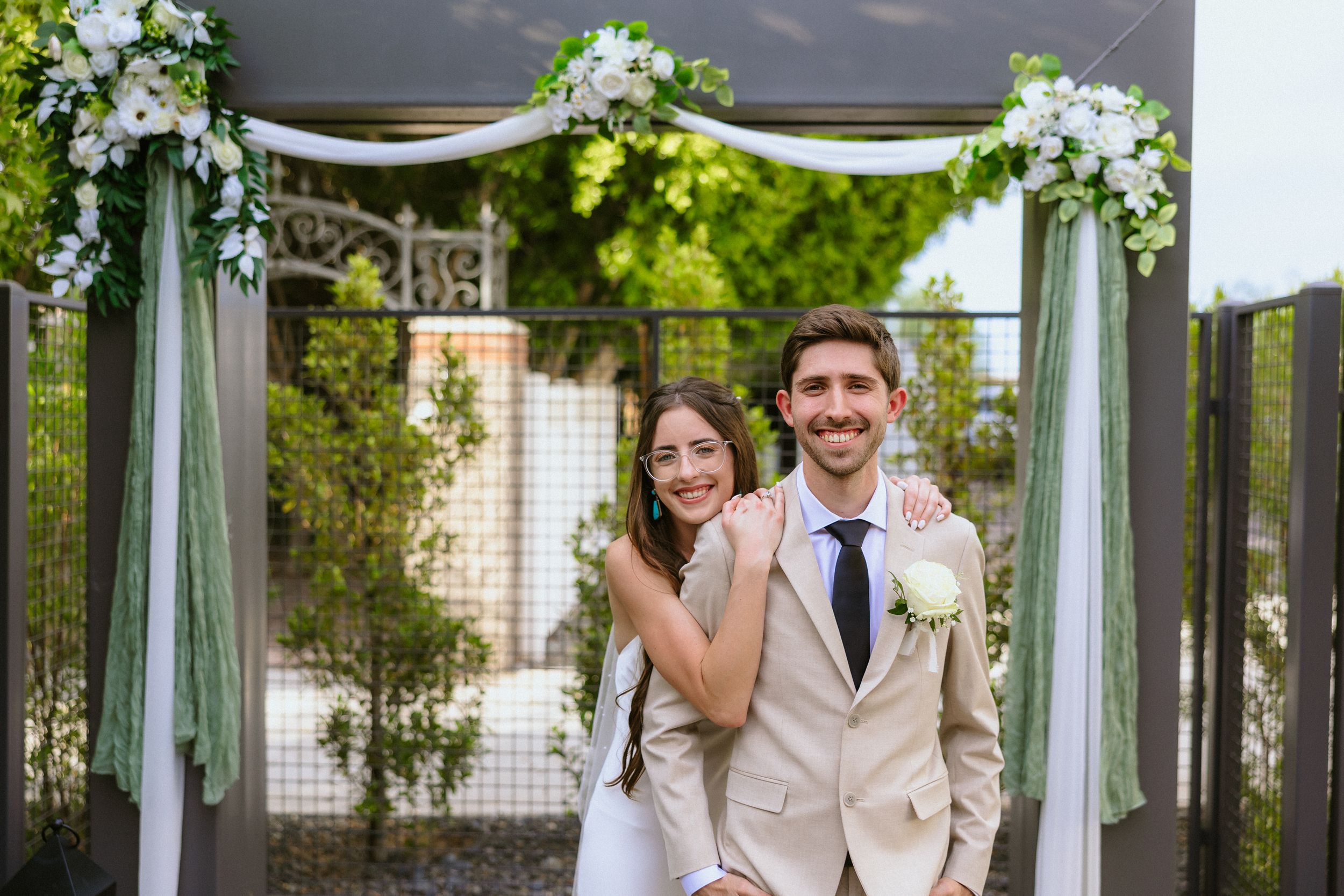 Happy couple posing under a floral wedding arch with white flowers and green draping outdoors.