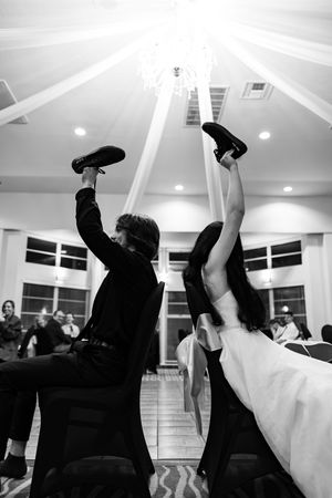 Black and white photo of bride and groom raising their shoes during a fun wedding reception chair dance.
