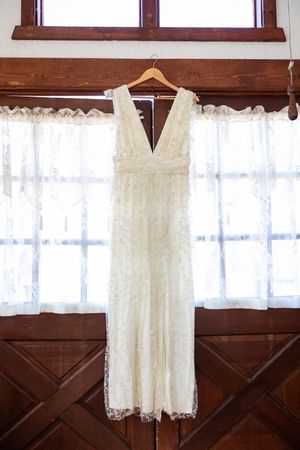 Elegant white lace wedding dress hanging on a wooden hanger in front of lace curtained windows.