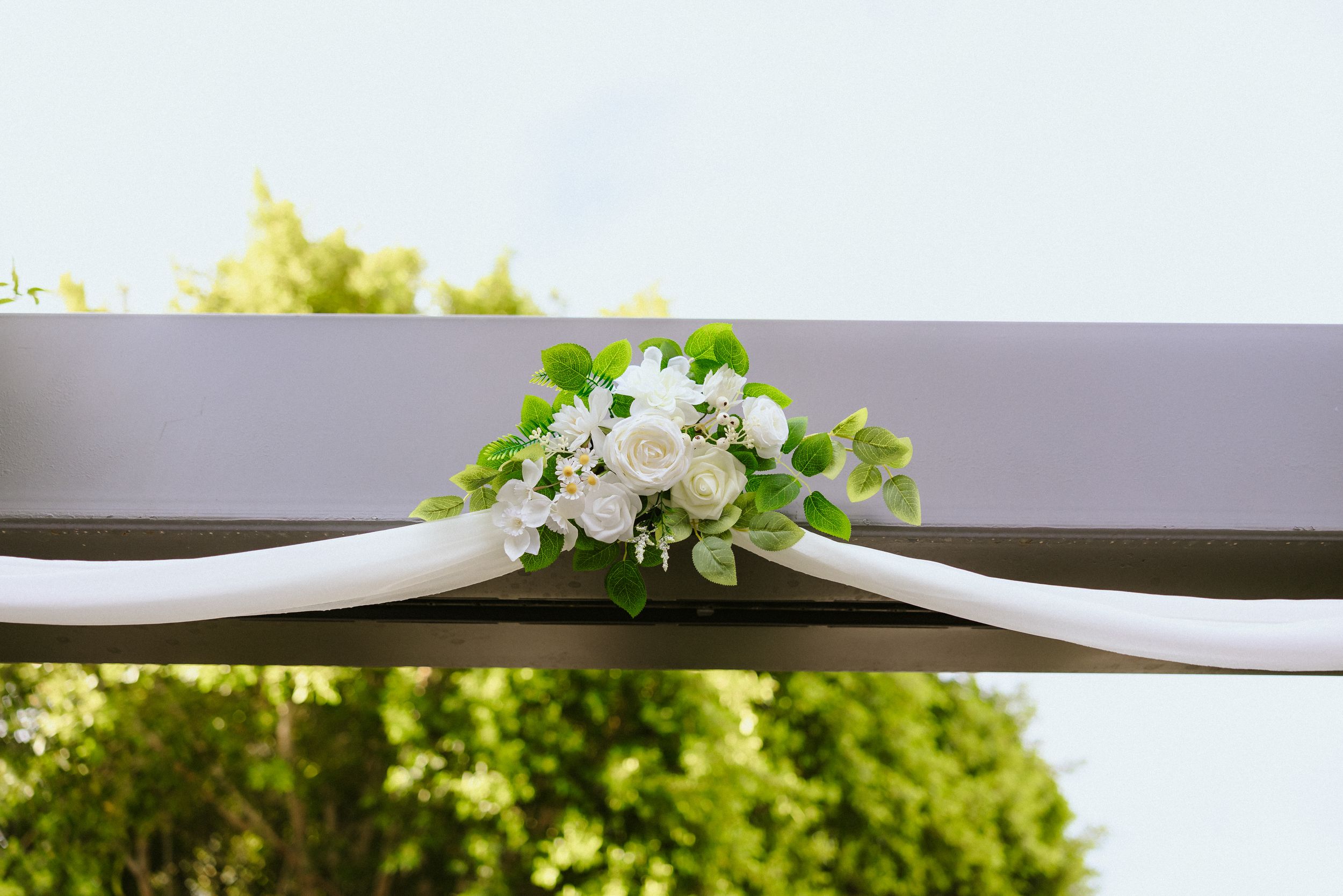 White roses and greenery floral arrangement on a decorated wedding arch with white draping outdoors.