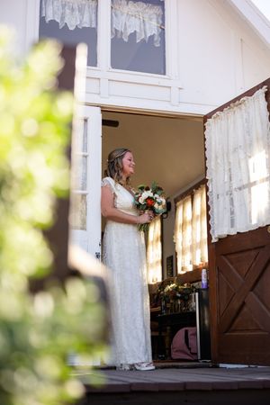 Bride in white lace gown holding colorful bouquet stands smiling in doorway of white building on wedding day.