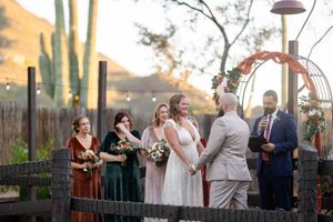 Outdoor desert wedding ceremony with bride, groom, officiant, and bridesmaids in green and rust dresses near saguaro cacti.