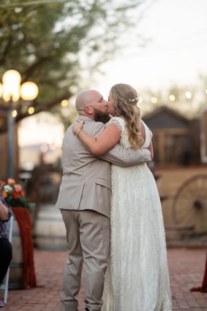 Bride and groom share their first kiss at an outdoor wedding ceremony with string lights glowing at dusk.