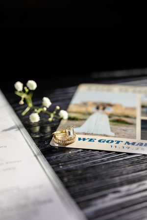 Gold wedding rings beside a We Got Married announcement card, white flowers, and a bridal photo on dark wood.