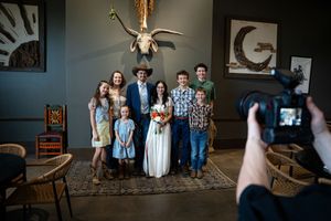 Wedding couple poses with family group indoors while photographer captures the joyful celebration moment.