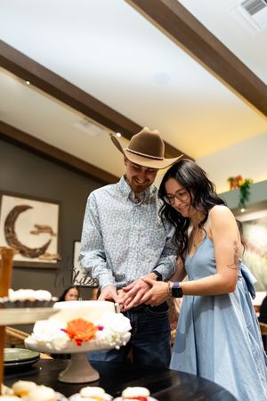 Couple cutting a white floral wedding cake together, man wearing cowboy hat and woman in blue dress.