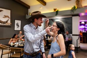 A man in a cowboy hat feeds a woman in a blue dress at a lively restaurant, sharing a playful romantic moment.