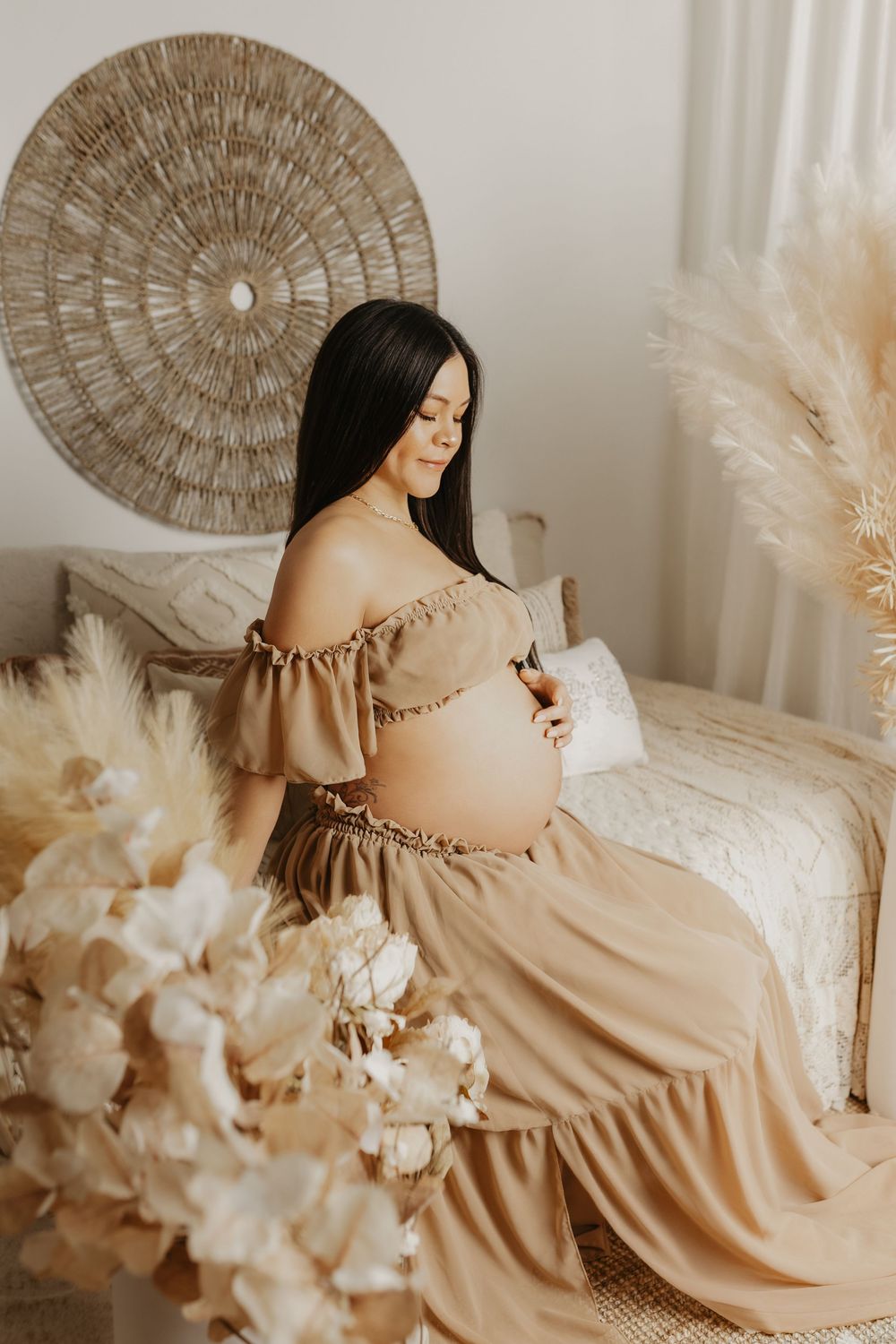 Maternity photoshoot featuring a flowing beige dress and boho decor with pampas grass and woven wall art.