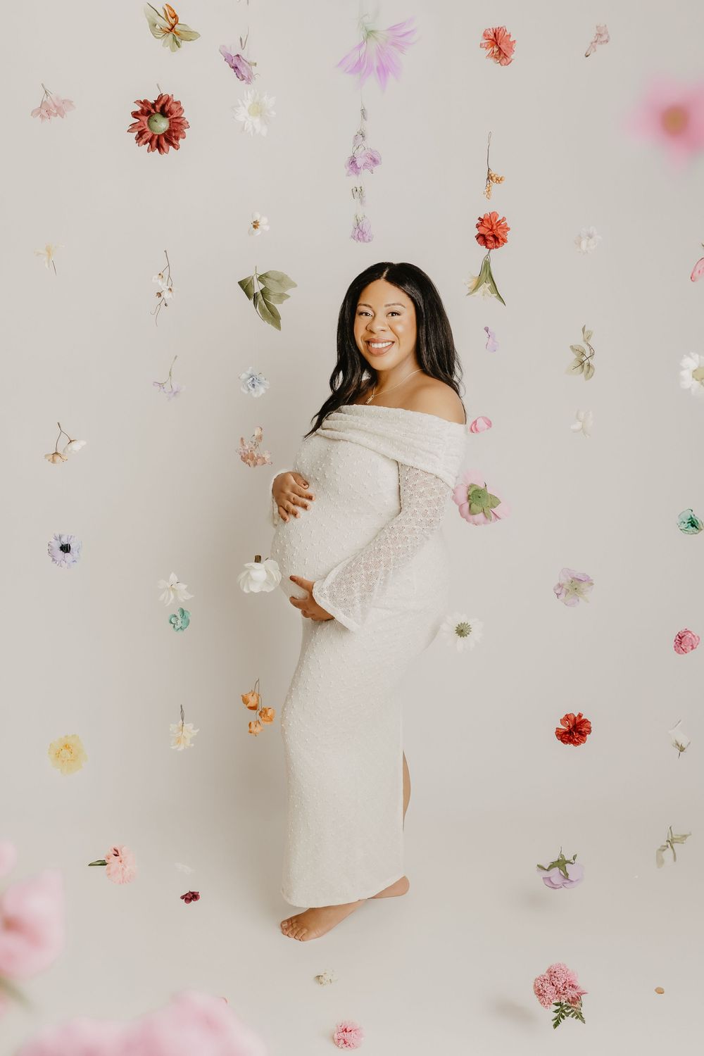 Woman in white dress poses joyfully against light background with floating colorful dried flowers.