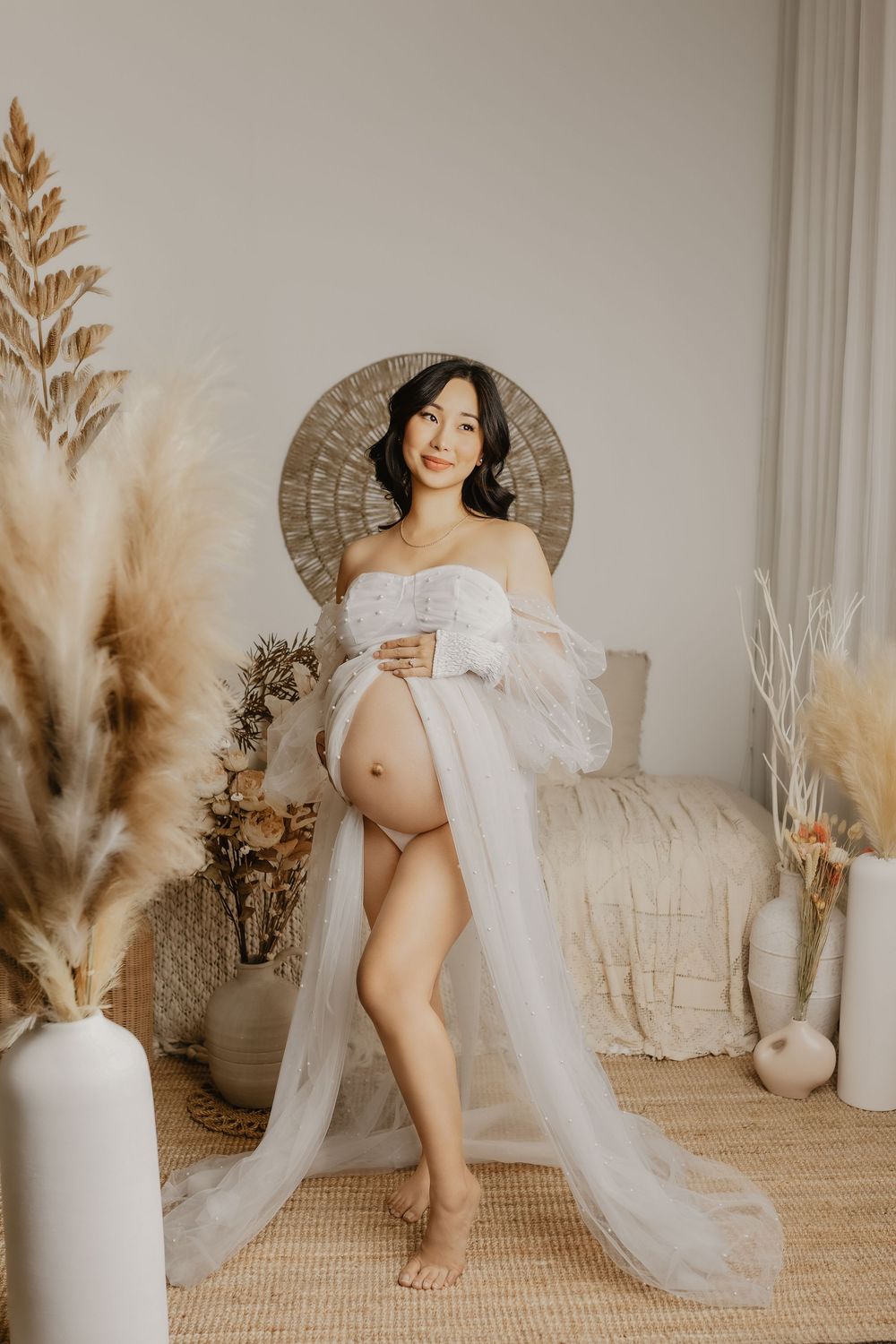 Artistic maternity photo featuring white fabric draping and pampas grass decor in a neutral studio setting.