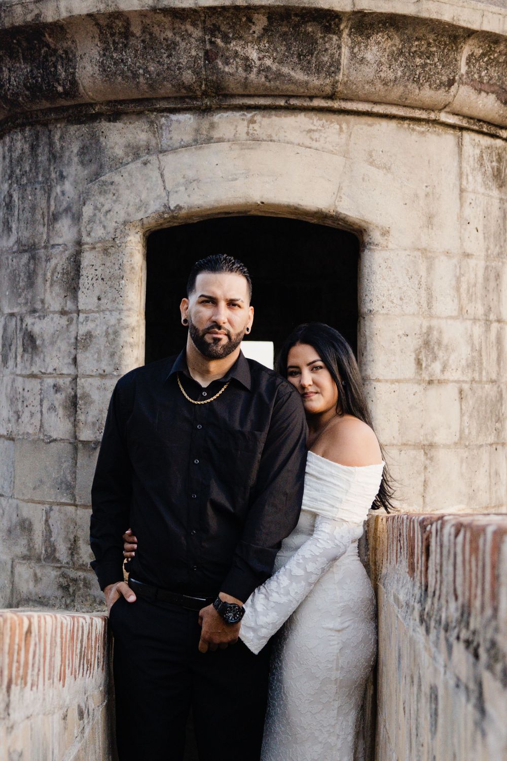 Couple in formal attire posing against a stone wall, man in black shirt and woman in white off-shoulder gown.