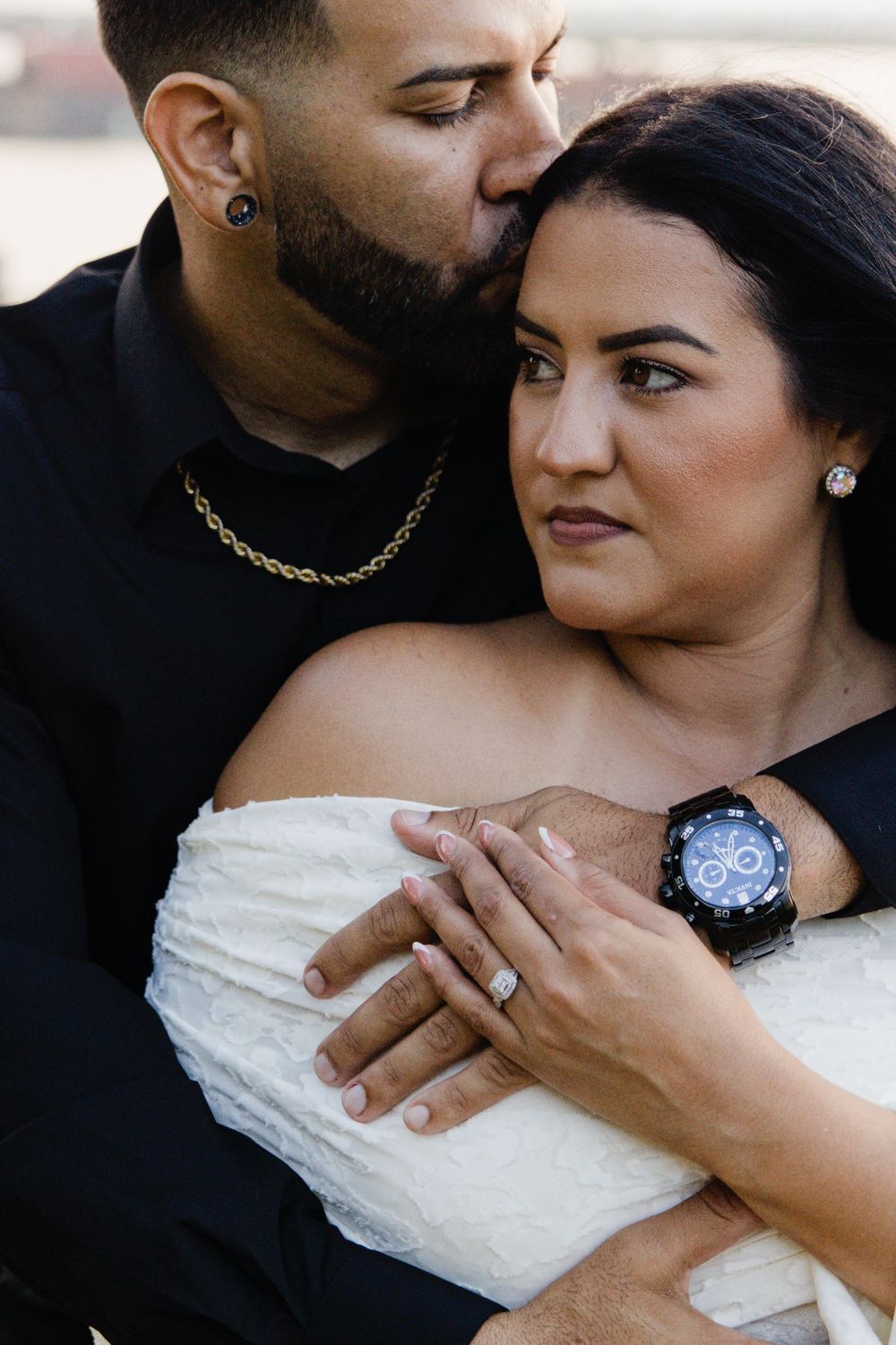 A man in a black shirt kisses a woman in a white off-shoulder dress while embracing her, showing her engagement ring.