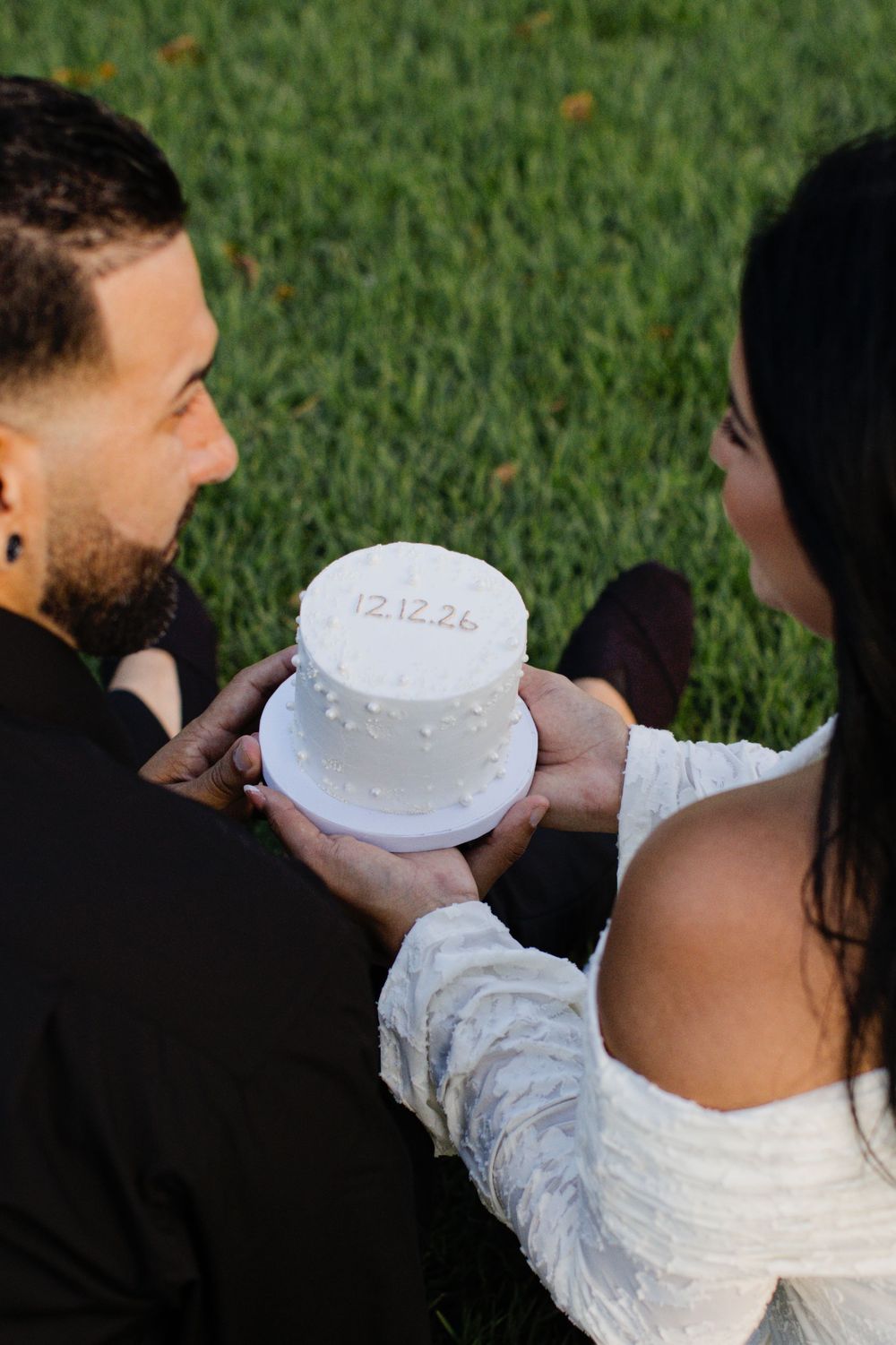 Couple holding a white wedding cake inscribed with date 12.12.26 while sitting on green grass.