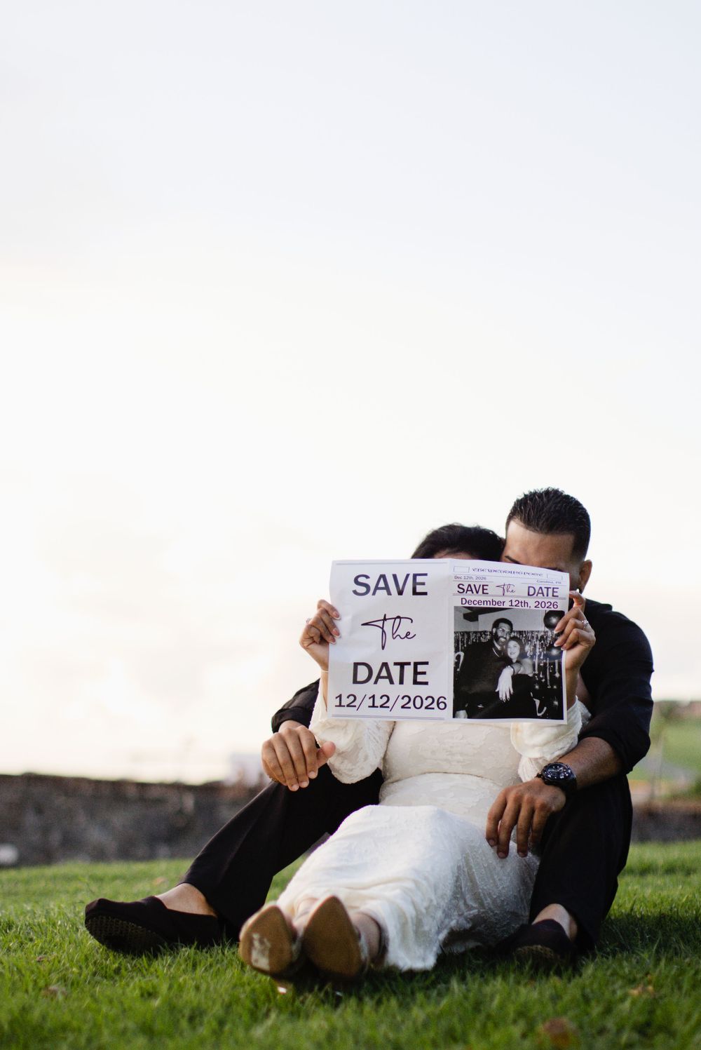 Couple sitting on grass holding a Save the Date sign for 12/12/2026, faces hidden behind the card.