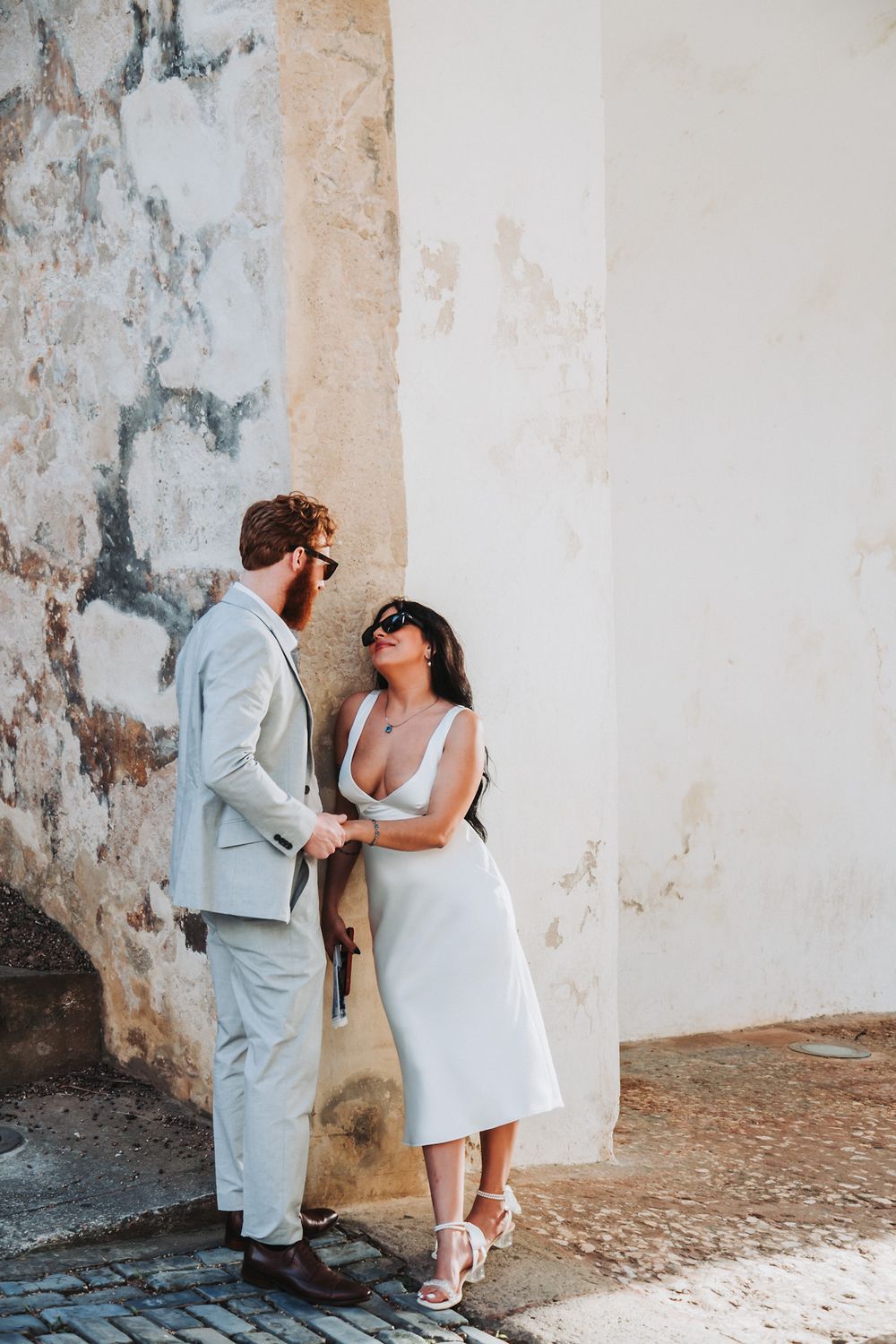 A romantic couple in light colored outfits poses against a weathered wall.