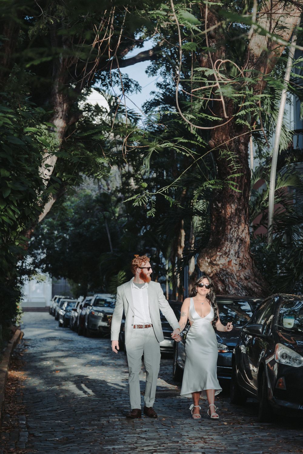 A couple in formal attire walks hand in hand down a tree-lined street.