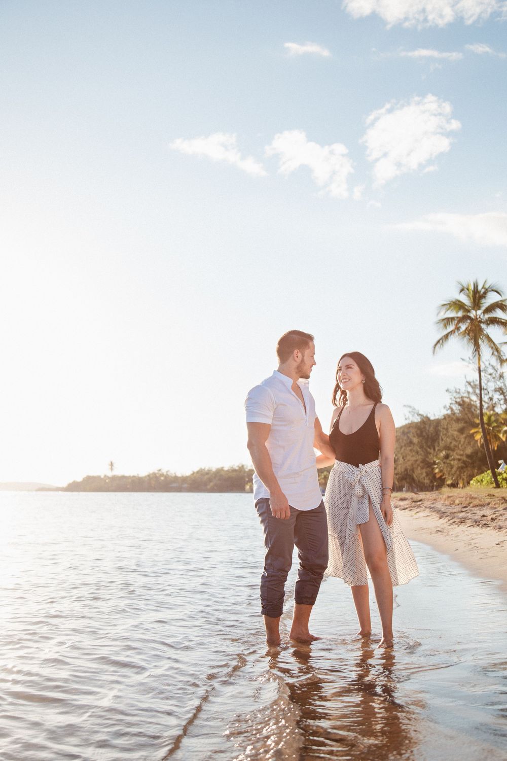 Couple standing barefoot in shallow ocean water on a tropical beach at golden hour with palm trees in background.