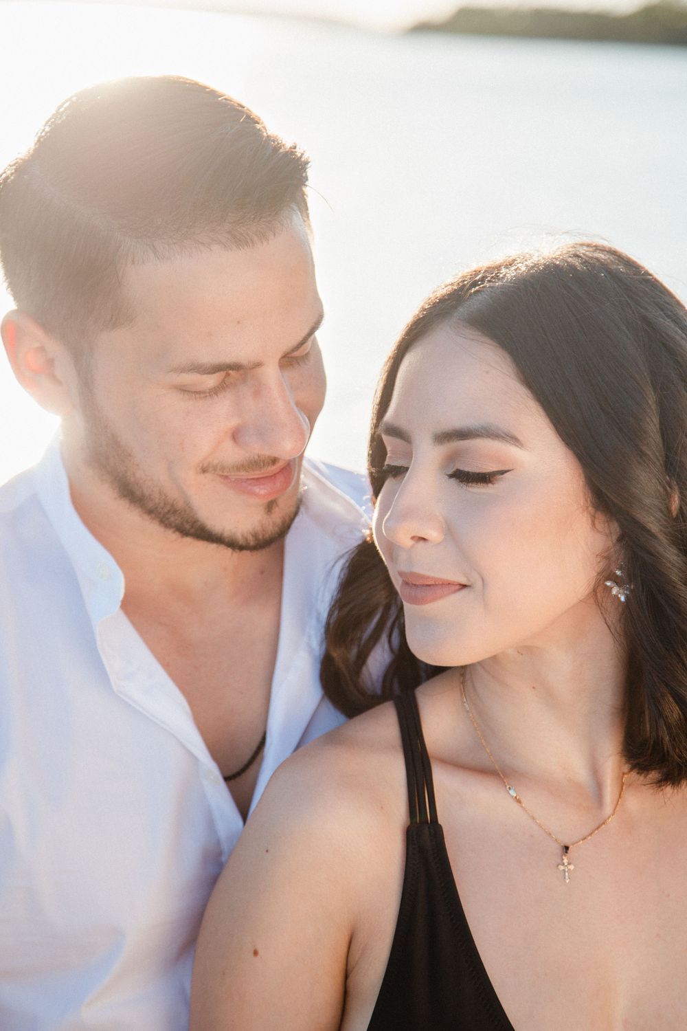 Romantic couple sharing a tender moment at sunset by the water, woman wearing a cross necklace.