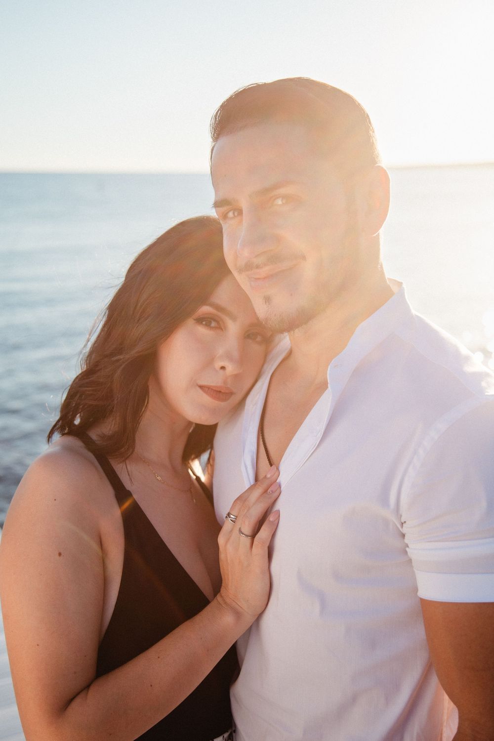 Romantic couple embracing at sunset by the ocean, woman in black dress, man in white shirt with golden backlight.