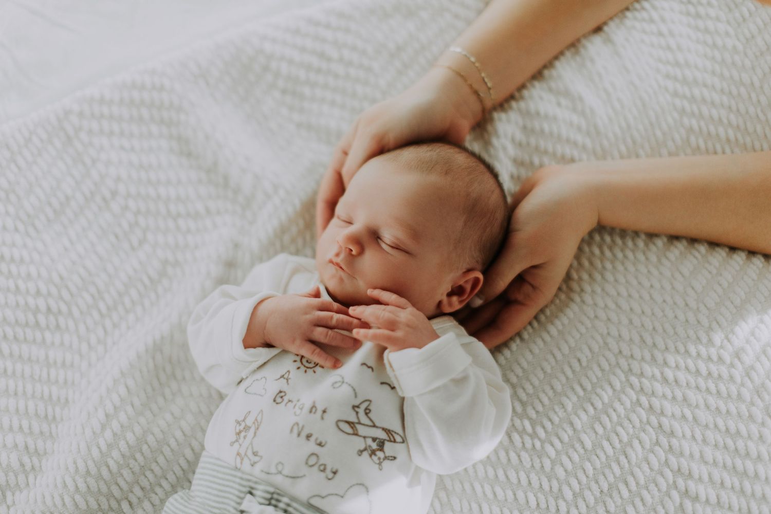 A sleeping baby in white clothing lies on textured bedding while a gentle hand touches their face.