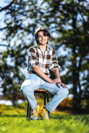 Casual portrait of person in plaid shirt and jeans sitting on stool in sunny outdoor setting.