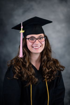 Graduate portrait in cap and gown with pink tassel against grey background.