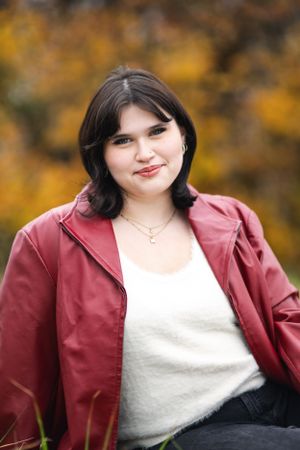 A person in a burgundy jacket poses outdoors with autumn foliage in the background.