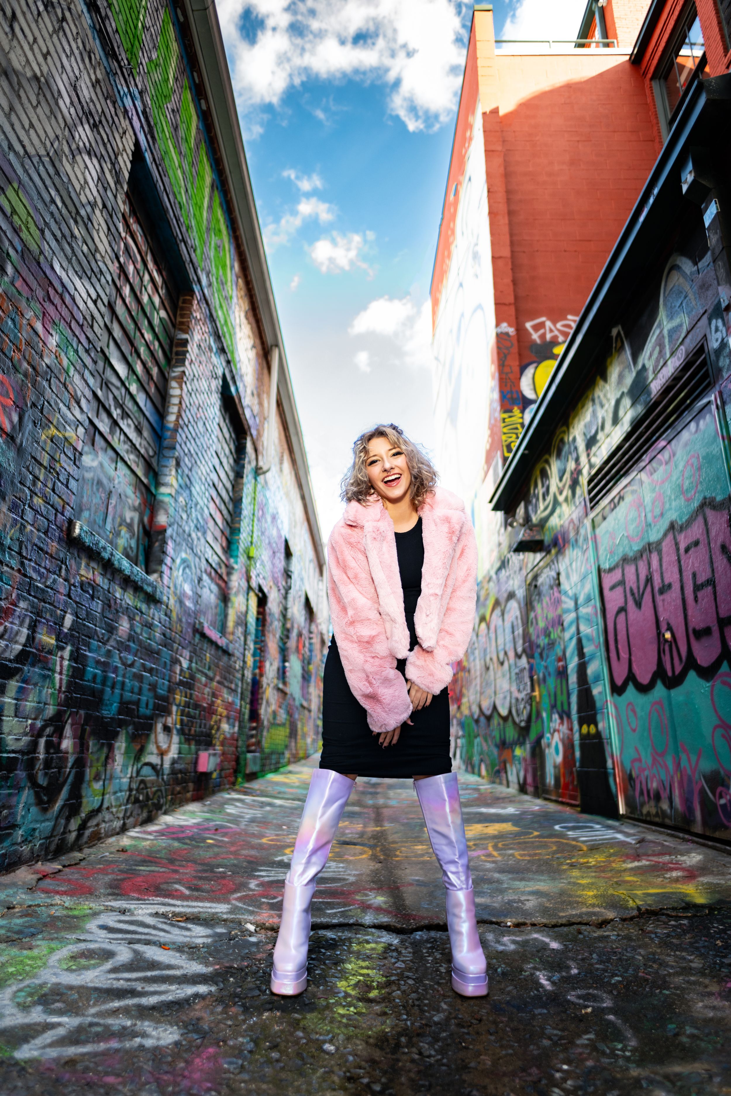 Fashion photoshoot in urban alley with graffiti walls featuring pink fur coat and silver boots styling.