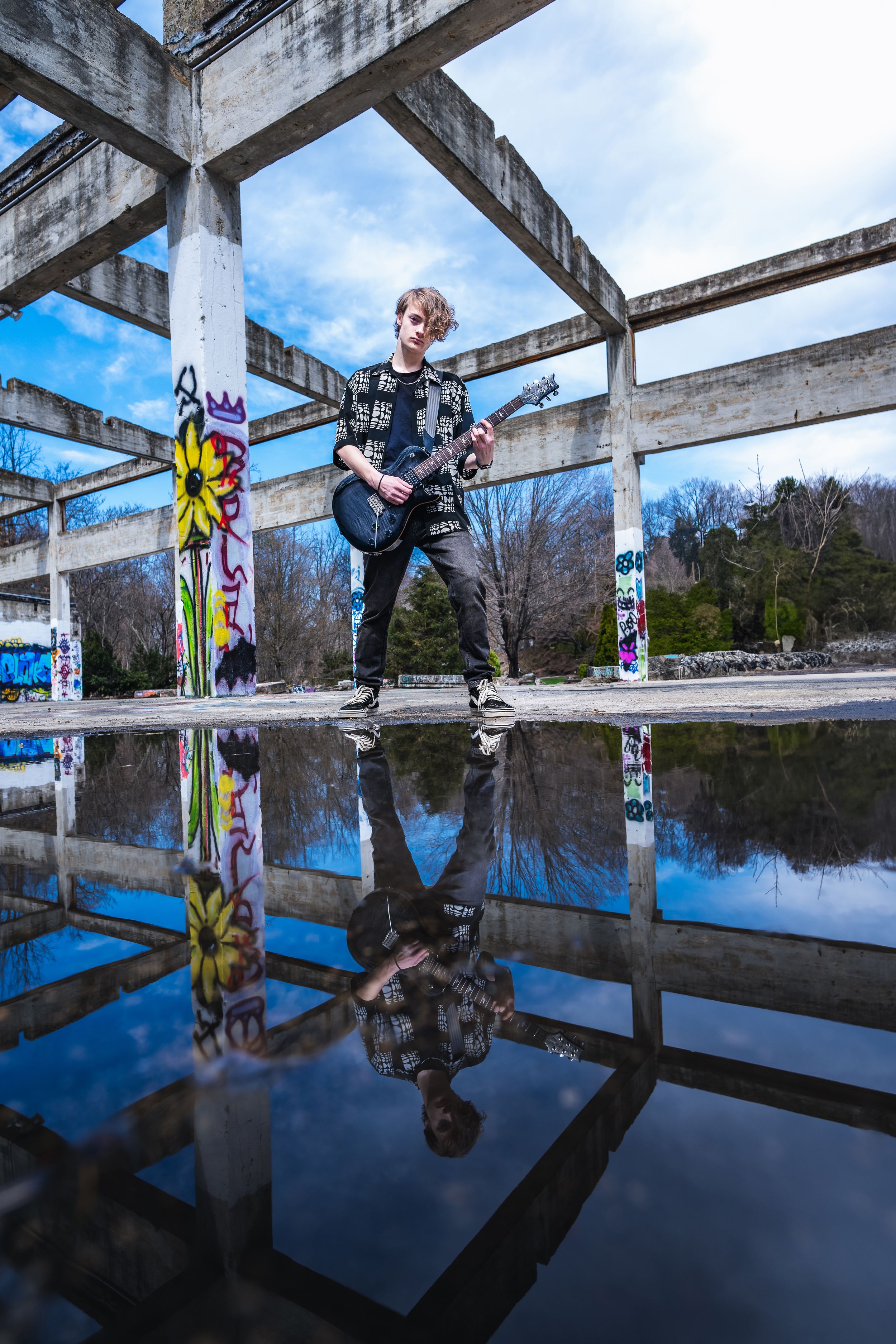 Young guitarist with electric guitar poses in abandoned concrete structure with graffiti, reflection visible in puddle below.