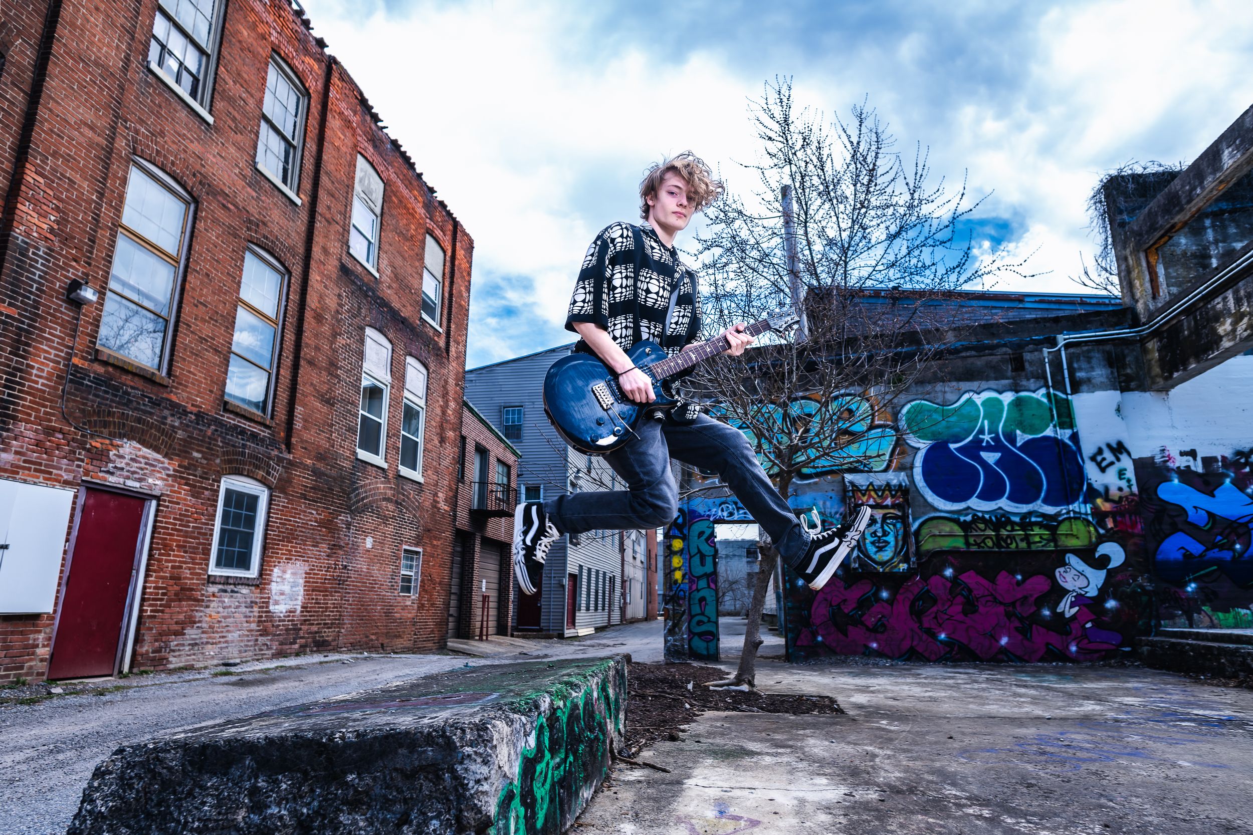 Young man jumping with guitar in urban alley with colorful graffiti walls.